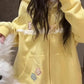 A person wearing a pastel yellow zip-up hoodie with white lace trim, a small cat graphic on the chest, and three pastel star patches on the front, taking a mirror selfie indoors.