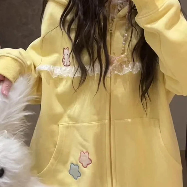 A person wearing a pastel yellow zip-up hoodie with white lace trim, a small cat graphic on the chest, and three pastel star patches on the front, taking a mirror selfie indoors.