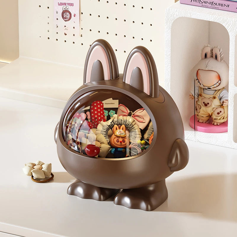 A brown rabbit-shaped decorative container with long upright ears, a clear oval window, and plush toys arranged inside, displayed on a white tabletop next to a small accessory piece and a shelf with a plush figure.