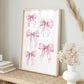 A framed art print featuring watercolor pink ribbon bows, styled on a woven table runner with dried grasses and a white vase in a neutral room setting.