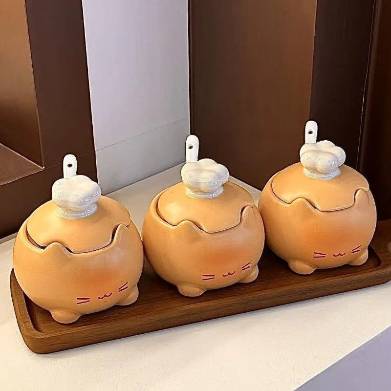 Three small ceramic kitchen containers with lid lids shaped like cartoon animals, arranged on a wooden tray against a kitchen background.