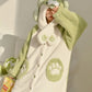 A person wearing a plush animal-themed hooded robe in pale green and cream, with paw-print details and a soft textured fabric, posed indoors against a light wall.