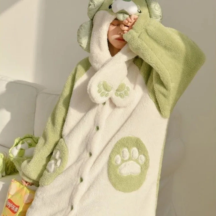 A person wearing a plush animal-themed hooded robe in pale green and cream, with paw-print details and a soft textured fabric, posed indoors against a light wall.
