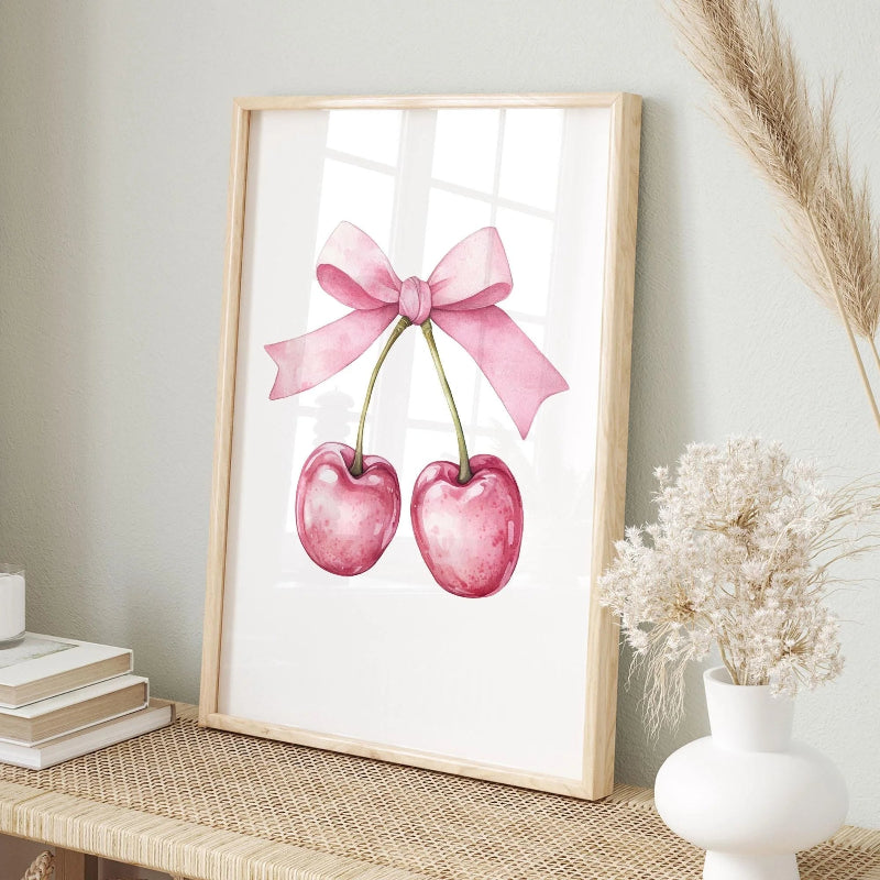 A framed watercolor art print featuring two pink cherries tied with a pink bow, displayed on a woven table in a styled room setting with books and dried flowers.
