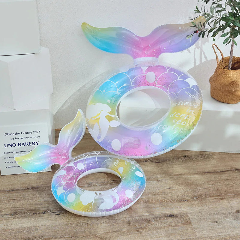 Colorful inflatable mermaid tail pool float on a wooden surface with a plant and bakery sign in the background.