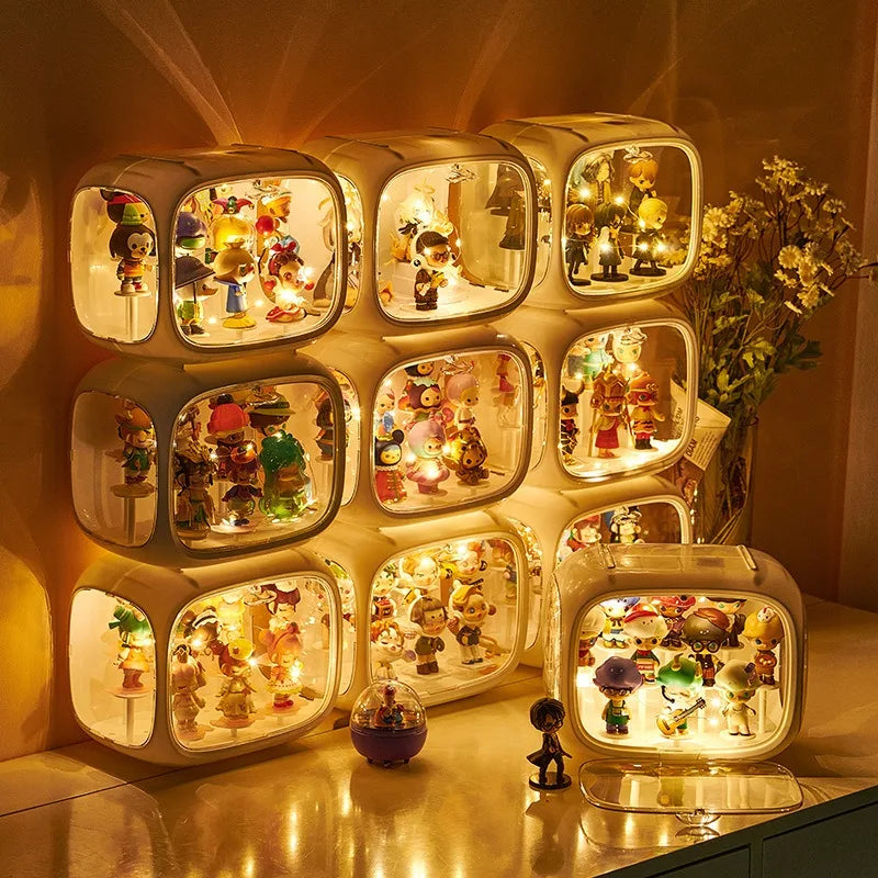 A set of nine illuminated display boxes arranged in a 3x3 grid on a tabletop, each lit from within to showcase small collectible figurines and toys. The boxes have rounded rectangular shapes with warm white lighting and are photographed in a cozy indoor setting with decorative flowers and a small figurine in the foreground.