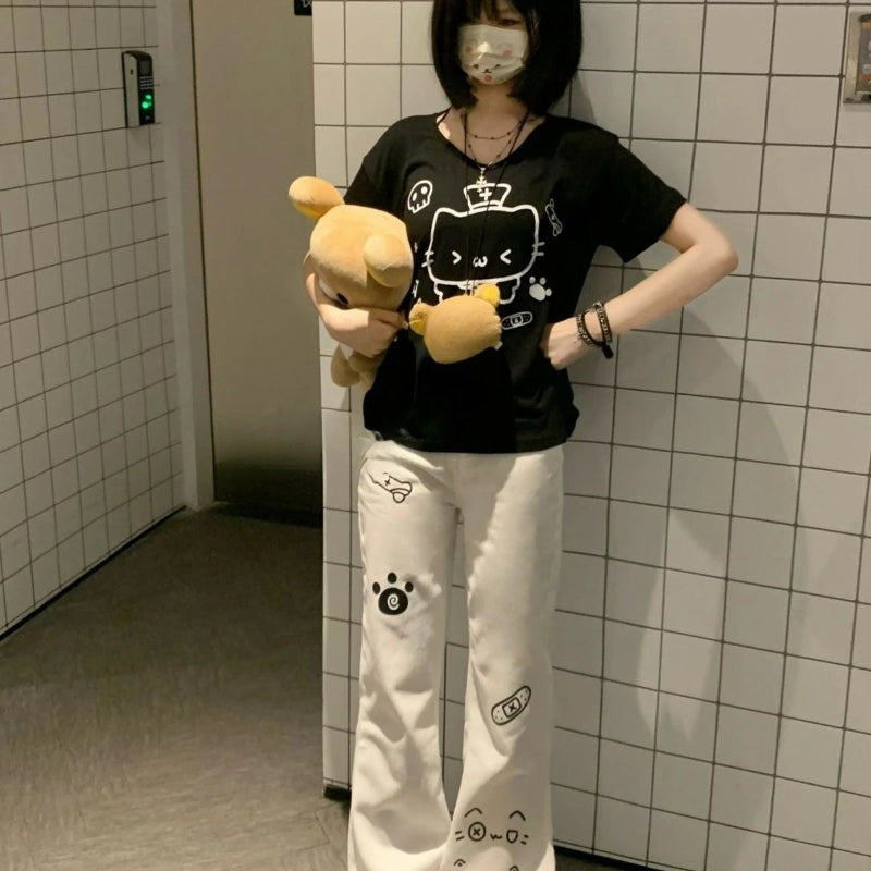 Person wearing a black t-shirt with a cartoon design and white pants with black paw prints, holding a plush toy, in a tiled room.