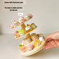 A three-tiered rotating display stand holding small pastel character figurines, shown with a hand holding the base and text callouts about stability and included base glue.