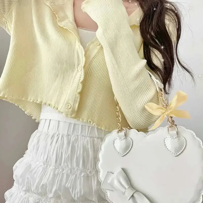 Person wearing a yellow cardigan and white skirt with a white handbag featuring heart designs and a bow.