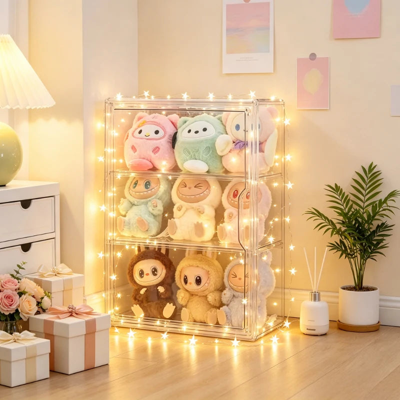 A clear acrylic three-tier display cabinet filled with pastel plush toys, decorated with warm string lights in a cozy, decorated room with a potted plant and gift boxes.