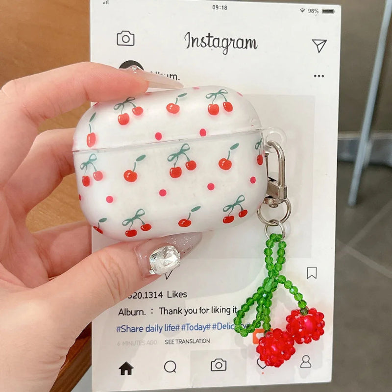 Hand holding a white airpods case with a cherry print design featuring a green beaded charm with red cherries.