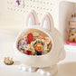 A white bunny-shaped decorative container with pink inner ears and a clear oval window, filled with colorful plush toys and accessories. The bunny sits on a white surface against a light cream wall with small pegs, with another plush figure visible in the background on the right.