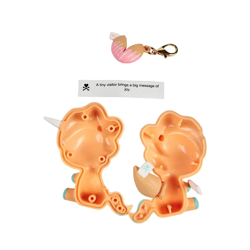A small novelty toy set on a white background, featuring two peach-colored plastic unicorn-shaped halves with small white horns, a pink striped egg-shaped piece with a gold clasp, and a small paper tag with text.