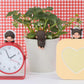 Small red alarm clock with cute figures, potted plant, and yellow heart-shaped object on a checkered background.