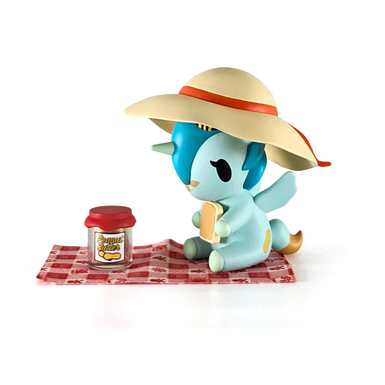 Toy figure of a blue unicorn with a sun hat and picnic blanket on a white background